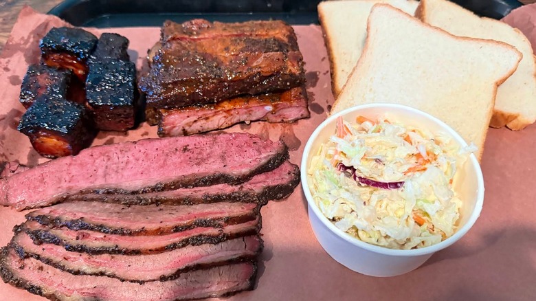 An overhead shot of a barbecue platter featuring sliced brisket with a pink smoke ring, burnt ends, and pork ribs, with sides of coleslaw and white bread