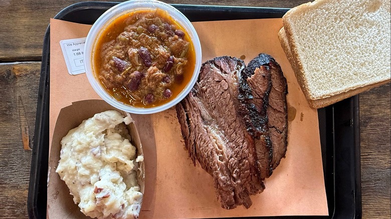 An overhead shot of a barbecue platter on a black tray, including sliced brisket with a dark, crusty bark, and a side of baked beans, potato salad, and two slices of white bread