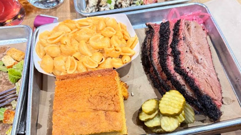 A barbecue platter on a metal tray, featuring thick slices of beef brisket with a dark, peppery bark, served with a large portion of cornbread, a side of macaroni and cheese, and sliced pickles
