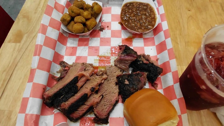 An overhead shot of a barbecue platter on red-and-white checkered paper, with sliced beef brisket that has a dark bark and pink smoke ring, baked beans, fried okra, and a slider bun