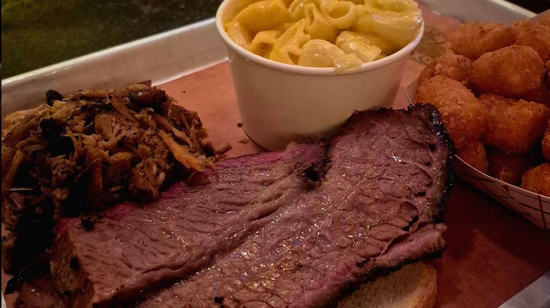An overhead shot of a barbecue platter on pink butcher paper, featuring thick slices of beef brisket with a noticeable smoke ring, pulled pork, a cup of macaroni and cheese, and crispy tater tots