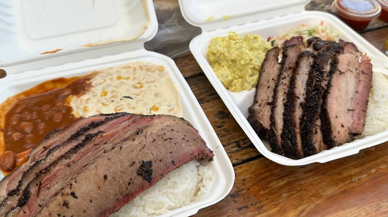 An overhead shot of two barbecue takeout containers on a wooden table, where both containers have thick slices of beef brisket with a dark, peppery bark, one container is served with a side of baked beans and creamed corn, and the other container is served with potato salad and coleslaw