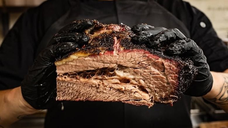 A person in black gloves and a black apron holding a large, sliced beef brisket with a dark, peppery bark, a distinct pink smoke ring, and a juicy, marbled interior