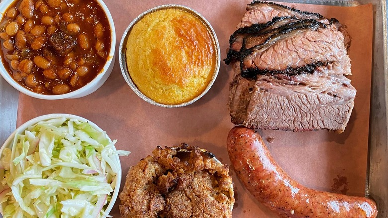 An overhead shot of a barbecue platter on pink butcher paper, featuring thick slices of beef brisket, sausage links, baked beans, coleslaw, cornbread, and a fruit cobbler