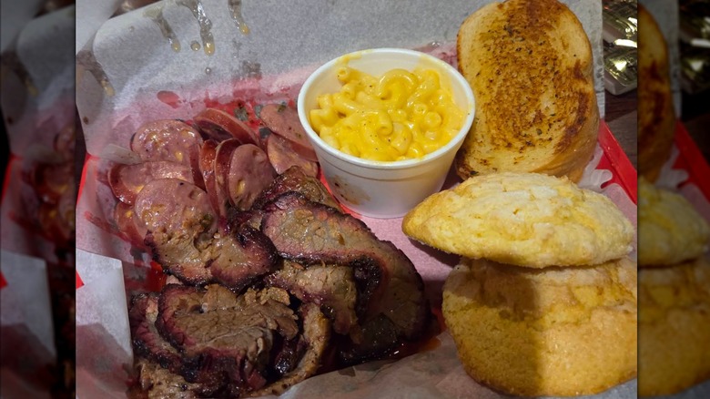 An overhead shot of a barbecue platter featuring sliced beef brisket with a pink smoke ring, a pile of sliced sausage, a cup of macaroni and cheese, a piece of toasted white bread, and two large cornbread muffins