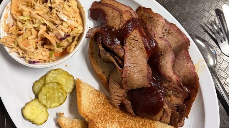 A white plate with sliced brisket, cole slaw, pickles, and toasted white bread