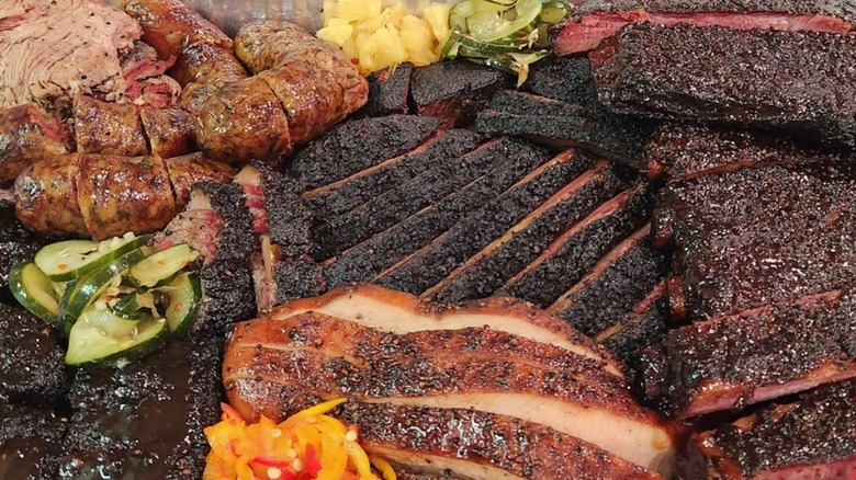 An overhead shot of a Texas-style barbecue platter featuring sliced beef brisket, pork ribs, sausage links, and pulled pork, garnished with pickled cucumbers and onions