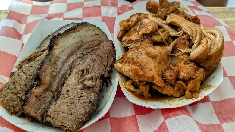An overhead shot of two white paper boats on red-and-white checkered paper, featuring thick slices of beef brisket with a dark bark and a side of pulled chicken or pork in a savory sauce