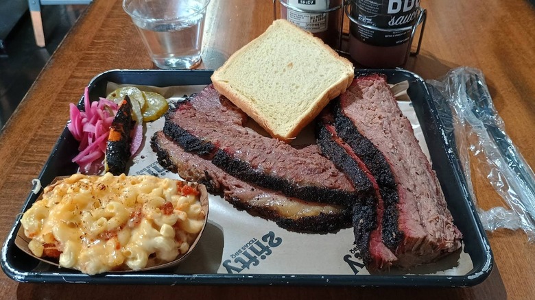 An overhead shot of a barbecue platter on a black tray, with thick slices of beef brisket, a side of macaroni and cheese, pickled red onions, pickles, and a slice of white bread