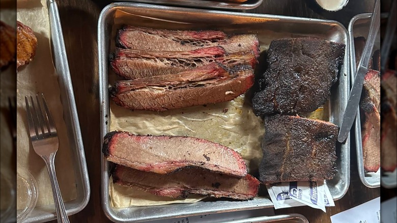 A metal tray with slices of smoked brisket with a dark, crusty bark and pink smoke ring and barbecue ribs