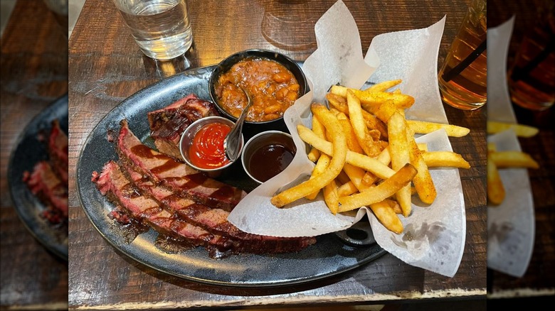 A barbecue plate with sliced brisket drizzled in sauce and a rack of ribs, a basket of seasoned fries, a bowl of baked beans, and small cups of barbecue and ketchup, on a wooden table