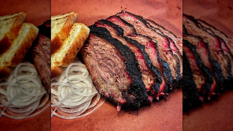 Slices of smoked brisket with a dark, charred bark and pink smoke ring, served with white bread and a side of thinly sliced raw onions