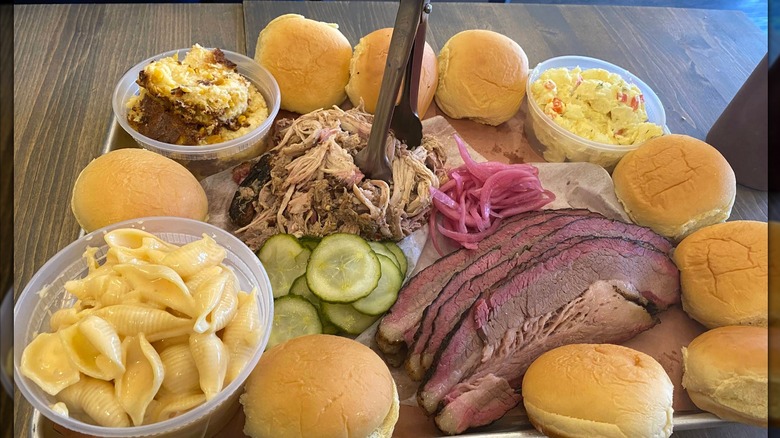 A barbecue platter with sliced brisket, soft slider buns, pickles, and pickled red onions, with sides of macaroni and cheese, potato salad, and cornbread