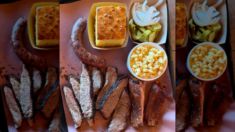 A barbecue tray lined with sliced brisket, pork ribs, and a grilled sausage, served with sides of macaroni and cheese, cornbread, pickles, and sliced onions