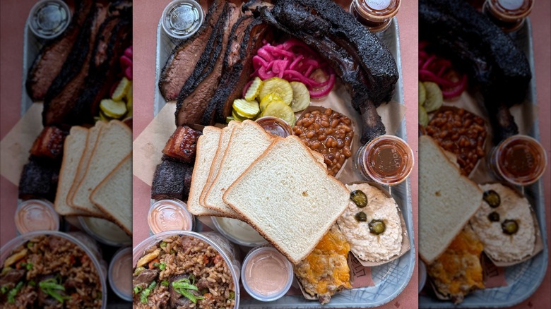A barbecue tray with sliced brisket, a beef rib, and chunks of pork, surrounded by white bread, pickles, and pickled red onions, with sides of baked beans, rice, mac and cheese, and dips, plus small cups of barbecue sauce