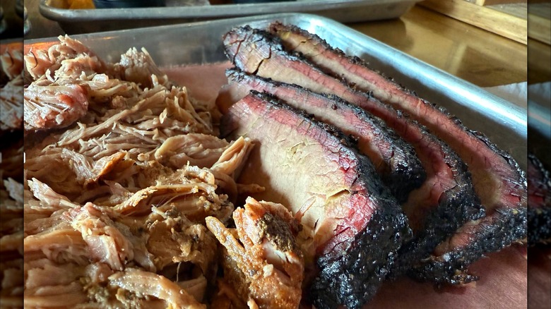 A metal tray lined with sliced brisket with a dark bark and pink smoke ring, served next to a pile of pulled pork