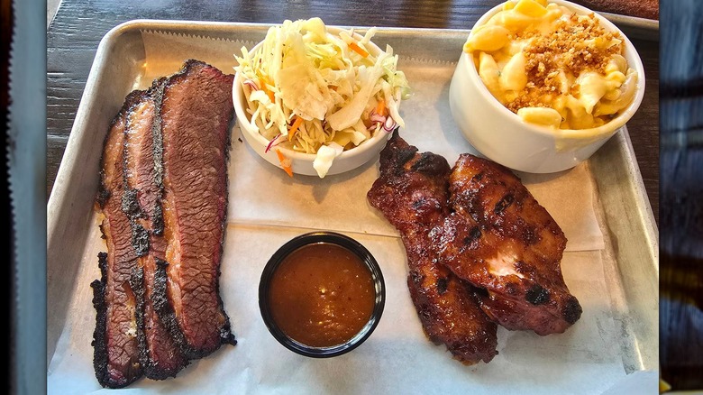 A metal tray with sliced brisket, barbecue chicken wings, and a barbecue cup of sauce, served with coleslaw and macaroni and cheese