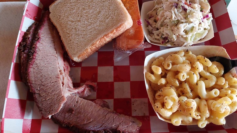 A red-and-white checkered tray with sliced brisket and a slice of white bread, served with sides of mac and cheese and coleslaw