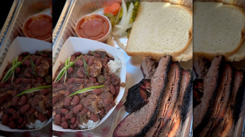 A foil takeout tray with sliced brisket and white bread, served with pickles, onions, a cup of sauce, and a side of beans over rice