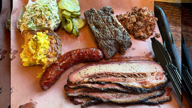 A barbecue tray with sliced brisket, a smoked sausage, and chopped pork, served with coleslaw, pickles, and sides of baked casserole and greens