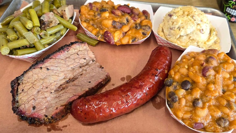 A barbecue tray with sliced brisket and a smoked sausage, served with sides of green beans, baked beans, and potato salad