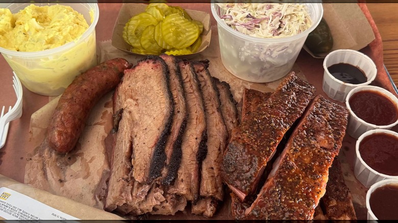 A barbecue tray with sliced brisket, pork ribs, and a smoked sausage, served with potato salad, coleslaw, pickles, and small cups of barbecue sauce
