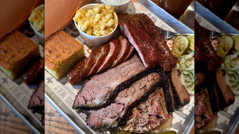 A barbecue tray with sliced brisket, pork ribs, sausage, and pulled pork, served with macaroni and cheese, coleslaw, cornbread, pickles, and a jalapeño