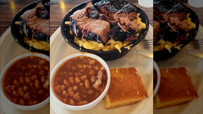 A bowl of macaroni and cheese topped with chopped brisket, served with baked beans and cornbread on the side