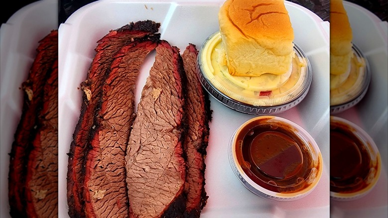 A takeout container with sliced brisket, served with potato salad, a slider bun, and a cup of barbecue sauce