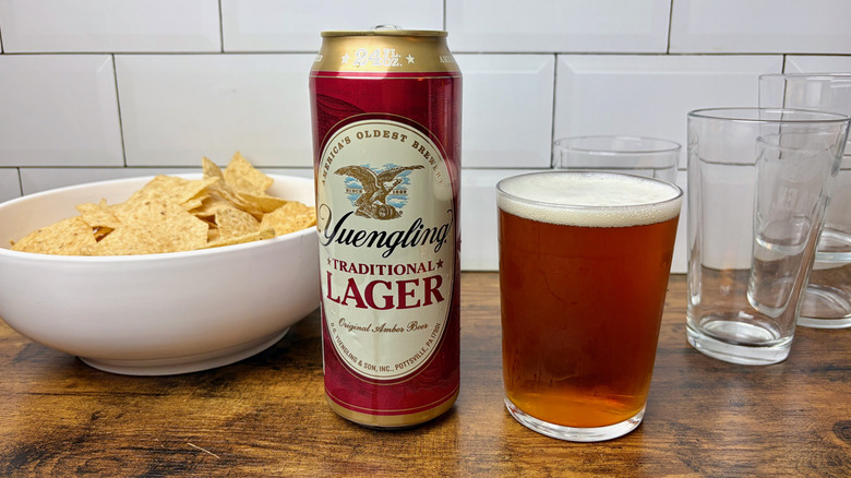 A tall boy can of Yuengling beer next to a poured glass and a bowl of tortilla chips