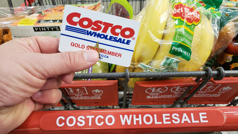 A person holding a Costco card in front of a Costco shopping cart. 
