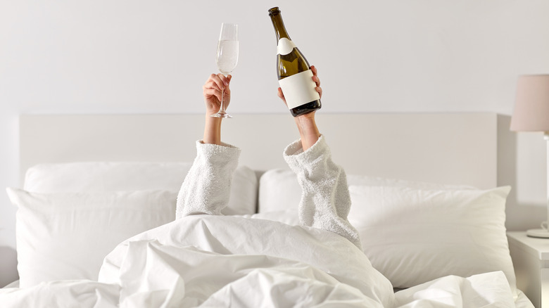 Guest in hotel bed holding up champagne bottle and glass