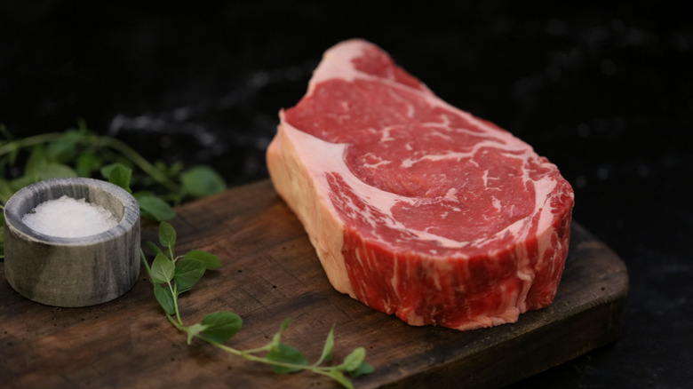 Raw thick ribeye on cutting board with salt and herbs.