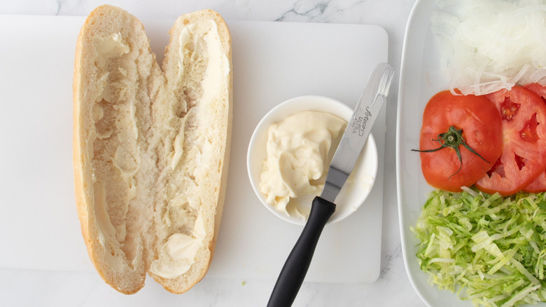 Hoagie roll spread with mayonnaise on cutting board next to plate with sliced tomatoes, shredded lettuce, and sliced onions