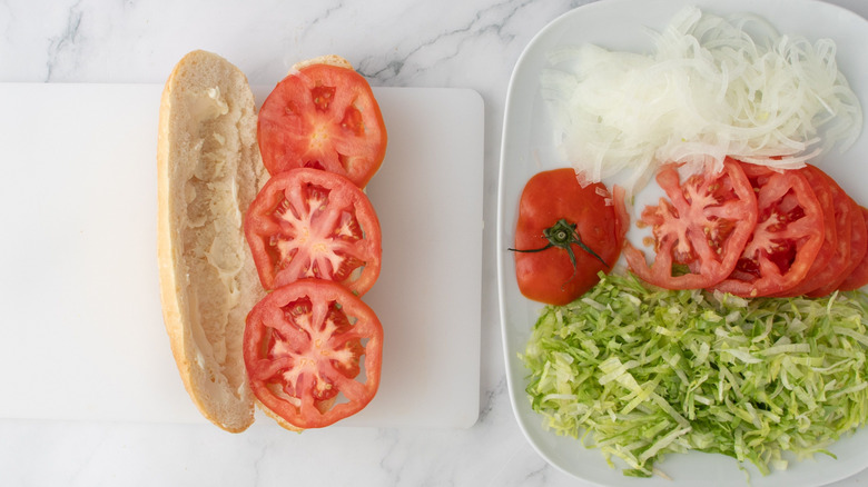 Hoagie roll with tomato slices next to plate with sliced tomatoes, sliced onions, and shredded lettuce