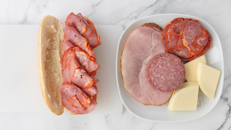 Hoagie sub topped with meat slices next to plate with cured meats and cheese