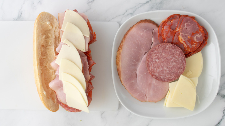 Hoagie topped with meats and cheese on cutting board next to plate with cured meat and cheese