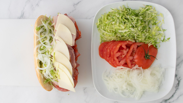 Sandwich with meat, cheese, lettuce, and onions next to plate with lettuce, sliced tomatoes, and sliced onions