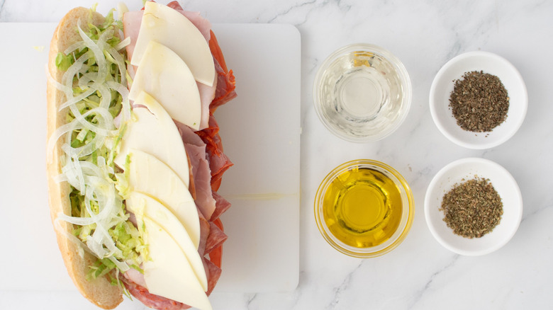 Sandwich with meat, cheese, lettuce, and onion next to small bowls of oil, vinegar, and seasoning