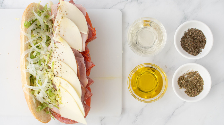 Sandwich with meat, cheese, lettuce, onion, and seasoning drizzled with oil next to small bowls of oil, vinegar, and seasonings