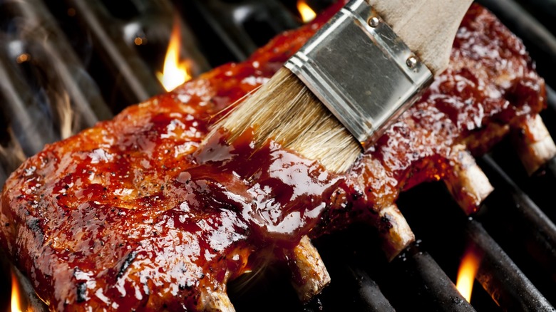 Barbecue brush spreading sauce on ribs set on flaming grill.