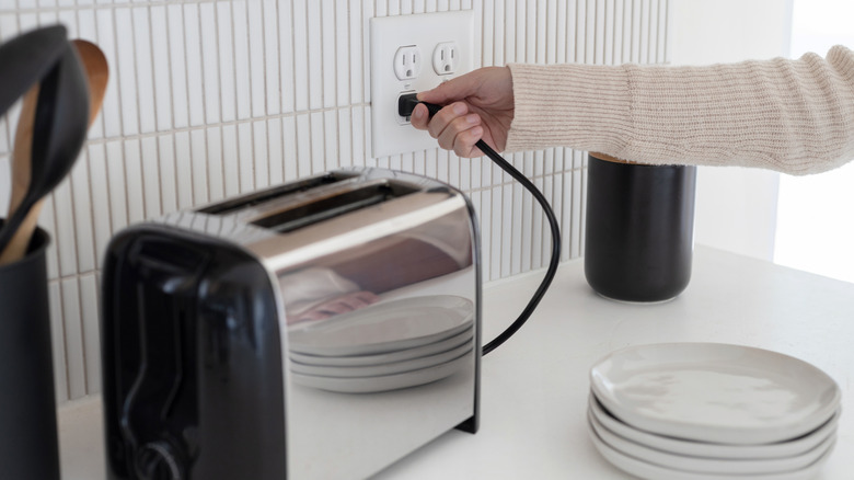 Someone plugs a toaster -- on a countertop with a stack of plates and a crock of kitchen spoons -- into a wall outlet.