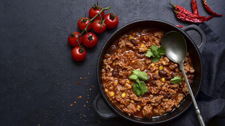 A pot of chili next to raw tomatoes and dried peppers