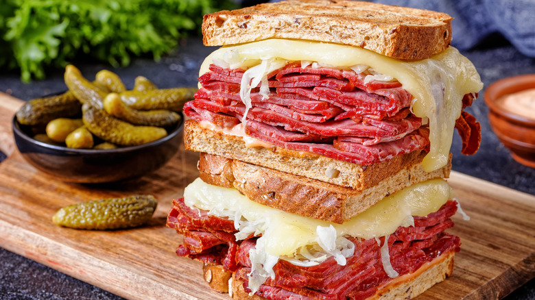 A corned beef sandwich next to a bowl of pickles on a wooden cutting board