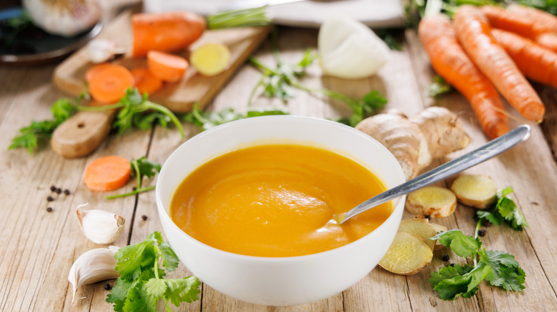 A white bowl of orange-hued soup surrounded by raw veggies on a wooden table