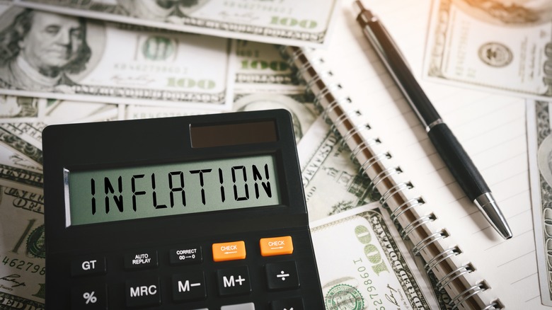 calculator reading "inflation" next to pen and adding book on pile of $100 bills