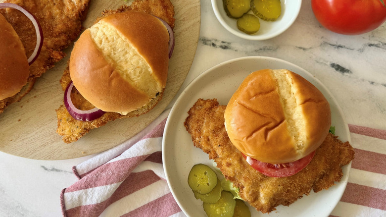 Hoosier pork tenderloin sandwiches on wooden board next to sandwich on plate