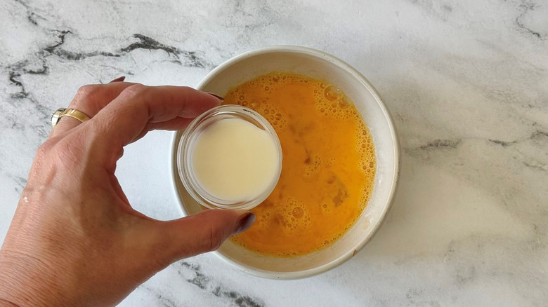 Hand holding small bowl of milk over bowl of beaten egg