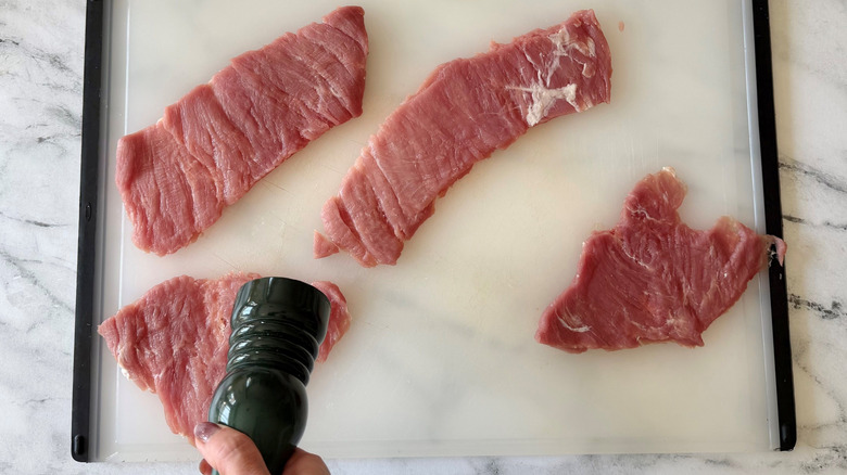 Hand holding pepper grinder over pork tenderloin pieces on cutting board