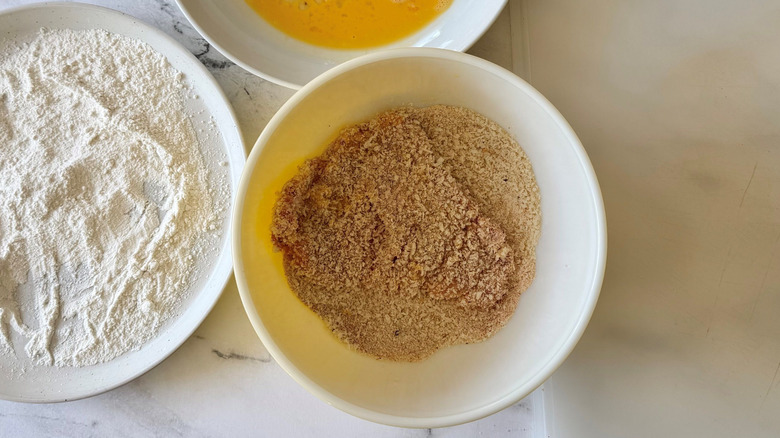Bowl of flour next to bowl of breadcrumbs with pork tenderloin in it